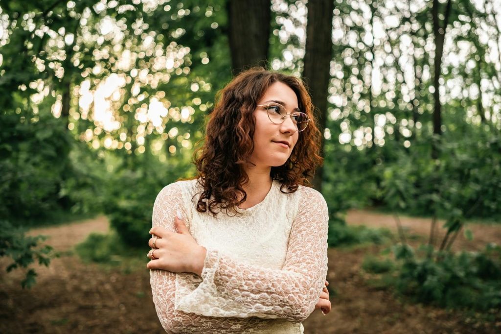 Woman with curly hair and glasses, wearing a lace dress, in a serene forest.