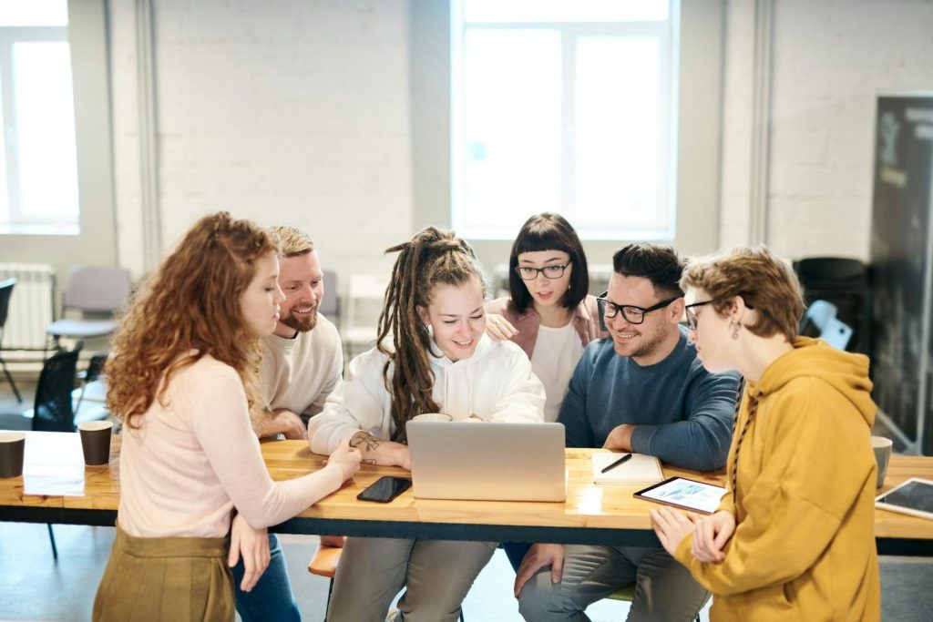 A diverse team of professionals collaborates happily around a laptop in a bright indoor setting.